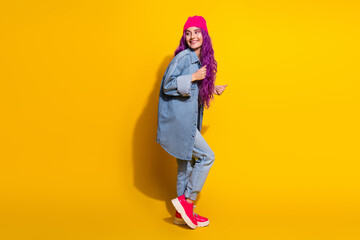 Trendy young woman with vibrant pink hair in denim outfit and beanie posing against a yellow background