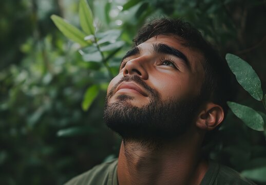 A young man with a beard looks upward amidst lush green foliage in a tranquil outdoor setting.