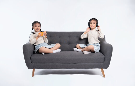 Two smiling girls on a couch, one with headphones and the other with a smart device, on a white background.