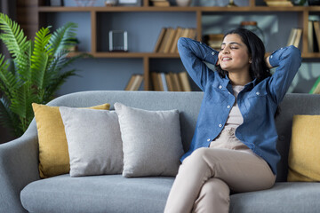 Obraz premium Relaxed young Indian woman in denim shirt sitting on sofa at home, hands behind head, eyes closed and resting
