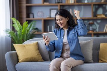 Obraz premium Happy young Indian woman in a shirt sitting on the couch at home, rejoicing at the news and result she received, reading it on a tablet she holds in her hand, celebrating victory