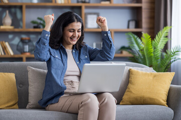 A young Muslim woman in casual clothes sits at home on the couch, holds a laptop on her lap, looks at the screen and rejoices at success and news, raising her arms up victoriously