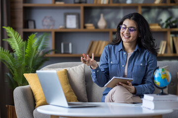 Indian young female teacher teaching remotely from home, sitting on sofa, holding notebook and...