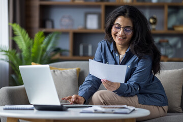 Smiling young Indian woman in casual clothes sitting on the couch at home and working remotely on a laptop, checking documents and bills