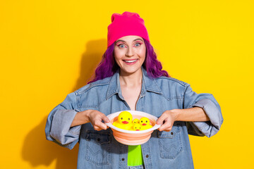 Smiling young woman holding toy ducks in a bowl against a vibrant yellow background, showcasing fun, casual style with pink hair