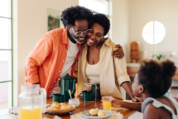 Mature parents hug each other at the breakfast table, enjoying a family meal with their daughter