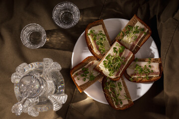 Slices of rye bread topped with salo and fresh microgreens, accompanied by vodka in crystal glasses, set on an elegant dark backdrop.