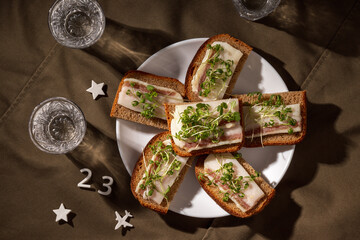 An artistic composition of rye bread with salo, fresh microgreens, and a glass of vodka on a dark...