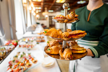 A person in a green outfit holds a tiered tray filled with assorted croissants. The blurred background features a buffet table, suggesting an event or catering service.
