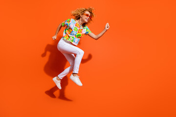 Energetic young woman in colorful t-shirt jumping excitedly against bright orange background, embodying happiness