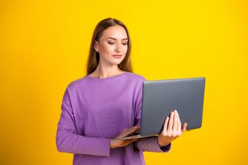 Fototapeta premium Young woman in a purple shirt holding a laptop confidently on a bright yellow background, exemplifying casual style