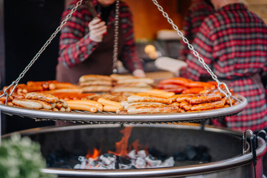 Grilled sausages cooking over an open fire on a hanging grill at an outdoor market. Vendors in plaid shirts prepare food in the background..