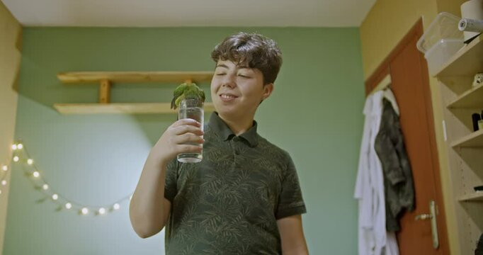 A young girl holds a glass of water for her pet green parrot to drink in her bedroom. The parrot perches on the glass and drinks. They enjoy their time together.