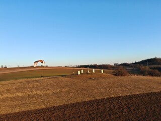 Obraz premium Motorized paraglider flying above a hill with a cross road between meadows, captured as aerial panorama showing sport aviation, landscape, and rural scenery