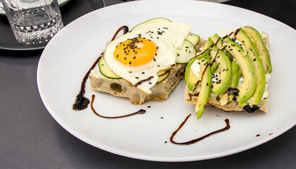 fancy avocado toast sandwich with cucumber and sunny side up egg, balsamic vinegar and sesame seed garnish served on a white plate from a restaurant in koper slovenia (mediterranean millenial food)