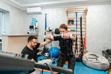 Obraz premium Professional physiotherapist working with beautiful young woman in EMS suit during special electro stimulation training or treatment. Modern and brightly lit studio.