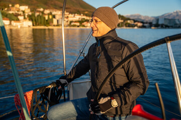 Obraz premium Young man captain standing at the helm and controls a sailboat during a journey by sea in winter time in Montenegro