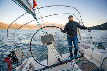 Young man captain standing at the helm and controls a sailboat during a journey by sea in winter...