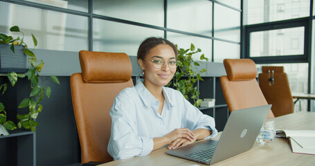  A professional young woman with glasses smiling confidently while sitting at a desk with a laptop in a modern, stylish office featuring plants and leather chairs