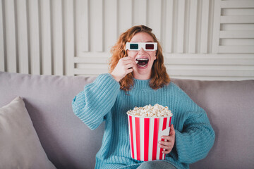 Obraz premium Young woman enjoying 3D movie at home while eating popcorn, wearing vibrant red-blue glasses and blue knitted sweater