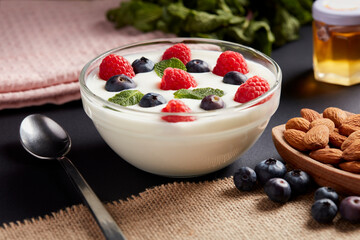 Un bol de yaourt crémeux garni de framboises, de myrtilles et de feuilles de menthe est présenté sur une surface sombre, accompagné d'amandes et d'une cuillère