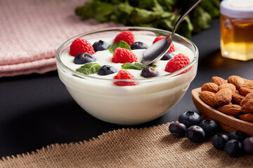 Un bol de yaourt crémeux garni de framboises, de myrtilles et de feuilles de menthe, surmonté d'une cuillère à café, est présenté à côté d'amandes dans un bol en bois