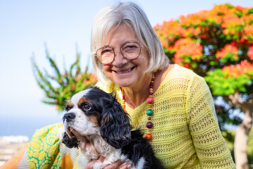 Portrait of senior smiling woman lying down in the meadow with her cavalier king Charles spaniel dog. Elderly lady and her best friend enjoying free relaxed moments