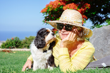 Smiling senior woman wearing hat and sunglasses lying outdoors in park with her cavalier king...