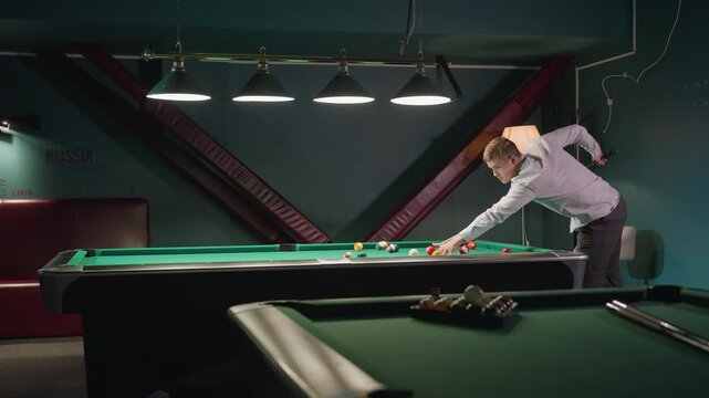 Billiards trainee in white shirt and grey trousers leans over green pool table, gripping cue stick with intense focus. Dark background as he strikes cue ball. Another pool table is visible