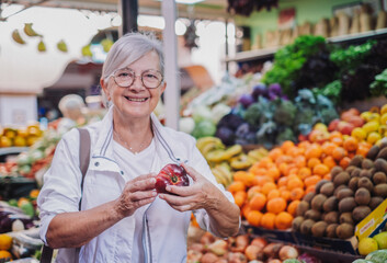 Senior woman holding a red tomato in hand while buying fresh fruit and vegetables at the market choosing from a large selection. Healthy eating, the concept of spending and consumerism