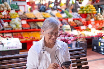 Smiling old mature modern senior woman using phone typing a message sitting on a bench at the fruits and vegetables local market.