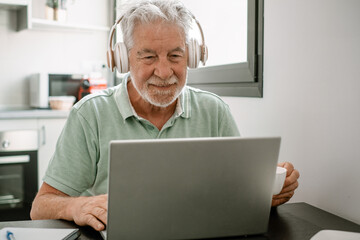 Smiling active modern white haired senior man with headphones sitting at home table using laptop in...