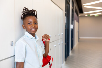Student standing in school hallway holding red backpack, smiling confidently, copy space