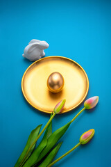 Golden easter egg displayed on a golden plate with a white ceramic bunny and flowers on a vibrant blue background, creating a luxurious and festive easter scene