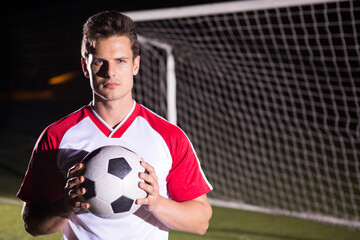 Soccer player holding ball confidently on field in front of goalpost at night, copy space