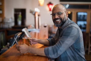 Smiling man enjoying beer and using smartphone at cozy bar counter, copy space