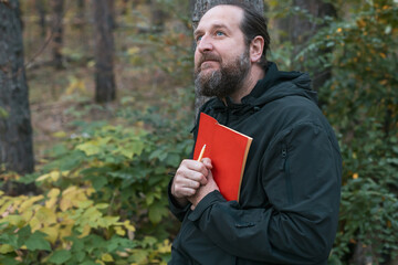 Fototapeta premium A man with a beard and a red book in his hand. He looks into the forest