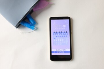 Smartphone with period calendar application and menstrual products on white table, flat lay