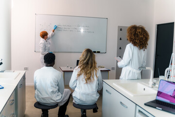 University professor writing chemical formulas on whiteboard, explaining lesson to students sitting...