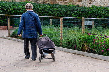 Senior citizen strolling with a shopping cart on a paved walkway next to a green wire fence and flowerbed