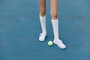 Female athlete showcasing sporty style in white knee high socks and sneakers on a bright blue court with a tennis ball, capturing an energetic and active mood