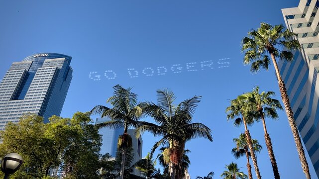 LOS ANGELES, CA, JUN 21, 2024: Downtown with the Deloitte building and "GO DODGERS" skywriting. Palm trees frame the scene against a bright blue sky