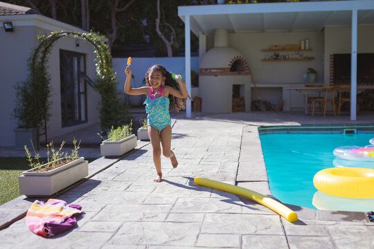 Young girl in swimsuit joyfully running by poolside holding popsicle, copy space