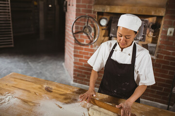 Female baker rolling dough on wooden table in rustic bakery kitchen, copy space