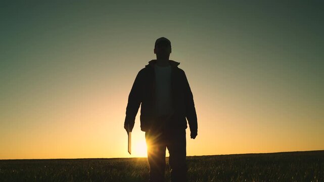 A man stands in a field at sunset, looking out into the distance. The sky is a deep blue, and the sun is setting, casting a warm glow over the landscape. The man is lost in thought