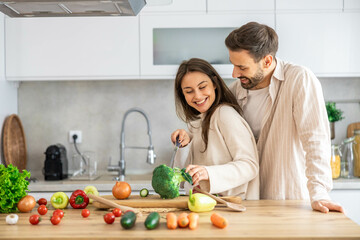 A couple is happily chopping broccoli together, surrounded by colorful vegetables in their cozy and modern kitchen setting.