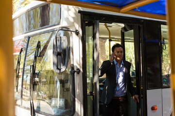 Businessman talking on smartphone while exiting bus at sunny urban stop, copy space