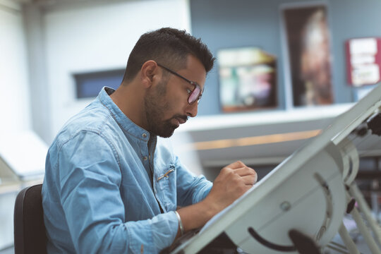 Designer in denim shirt sketching at drafting table in modern office
