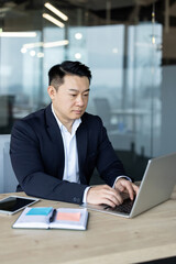 Vertical photo of an Asian young man in a suit sitting at a desk in an office and working on a laptop and documents