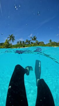 Underwater scene in Thoddoo, Maldives with a view of submerged legs in fins, clear turquoise water, and palm trees in the background.